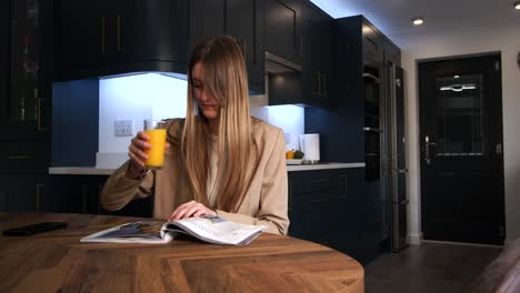 female-model-in-a-smart-jacket-drinking-from-a-glass-of-orange-juice-reading-a-magazine-and-looking-at-phone-at-a-kitchen-table-in-a-modern-kitchen