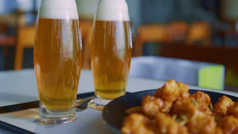 Dos-Vasos-De-Cerveza-Lager-Bebidas-Espumosas-Con-Un-Plato-De-Comida-Ssnackin-Un-Restaurante-Café-Fiesta-Con-Un-Camarero-En-El-Fondo