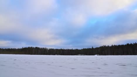 Winter-landscape-at-a-frozen-lake,-forest,-flying-clouds-in-a-blue-sky