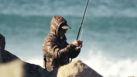 Fisher-preparing-fishing-rod-and-bait,-ocean-waves-in-background,-sunny-day
