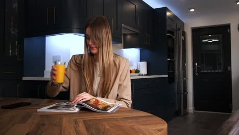female-model-in-a-smart-jacket-drinking-from-a-glass-of-orange-juice-reading-a-magazine-at-a-kitchen-table-in-a-modern-kitchen
