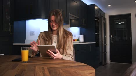 female-model-in-a-smart-jacket-drinking-from-a-glass-of-orange-juice-using-a-iPad,-tablet-at-a-kitchen-table-in-a-modern-kitchen