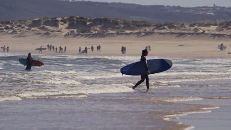 Surfer-walking-shallow-ocean-water-away-from-beach,-people-in-background