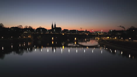 Low-angle-drone-panning-shot-with-a-reflection-of-the-Wettstein-bridge-in-the-Rhine-river-and-in-the-background-the-skyline-of-Basel-after-sunset