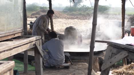 Male-Cooks-Seen-In-Rural-Punjab-Beside-Large-Cooking-Pan-And-Stirring-With-Steam-Rising-From-It,-Making-Gur-Jaggery-Slow-Motion