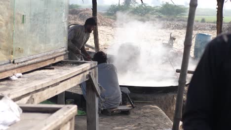 Male-Cooks-Seen-In-Rural-Punjab-Beside-Large-Cooking-Pan-With-Steam-Rising-From-It,-Making-Gur-Jaggery-Slow-Motion