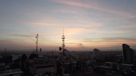 IN-THE-FOREGROUND-A-TELECOMMUNICATION-ANTENNA-IS-SEEN-IN-THE-BACKGROUND-VOLCANOES-ARE-SEEN-DURING-A-DAWN-IN-A-CITY