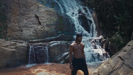 Drone-forward-moving-shot-of-a-black-male-standing-on-the-rock-in-front-of-jungle-waterfall-surrounded-by-lush-green-vegetation-at-daytime