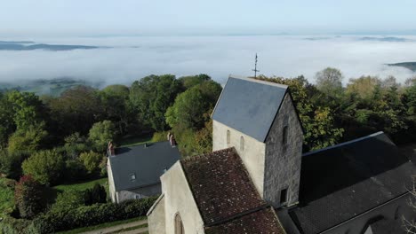 Drone-flying-over-church-of-Saint-Roch-in-Uchon-with-fog-shrouded-valley-in-background,-Saone-et-Loire-department-in-France