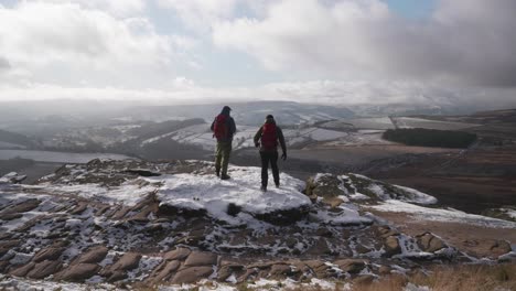 Two-men-standing-on-a-snowy-outcrop-admiring-a-beautiful-landscape-view-of-a-frozen,-wild,-rural-expanse-in-the-Peak-District