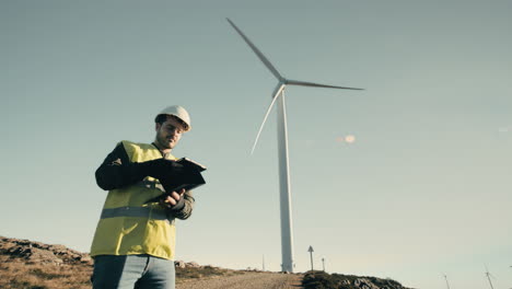 A-wide-shot-of-a-young-male-engineer-walking-towards-a-wind-turbine-and-then-using-technology-on-a-tablet-to-check-the-windmill,-emphasizing-the-role-of-innovation-in-the-production-of-clean-energy