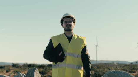 A-skilled-Caucasian-male-engineer-in-safety-gear-inspects-wind-turbines-while-walking-in-a-field-of-renewable-energy-generators-on-a-sunny-day,-emphasizing-the-significance-of-renewable-energy