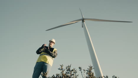 Advancing-renewable-energy,-an-engineer-uses-a-tablet-to-audit-wind-turbines-in-a-field-of-clean-energy-generators-on-a-sunny-day,-wearing-a-white-helmet-and-reflective-vest