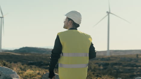 Walking-among-wind-turbines,-a-young-Caucasian-engineer-in-a-white-helmet-and-vest-represents-the-growth-of-clean-energy-for-a-greener-future