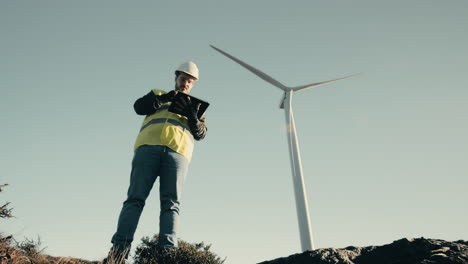 Ensuring-a-sustainable-future,-an-engineer-in-a-white-helmet-and-reflective-vest-uses-a-tablet-to-audit-wind-turbines-in-a-field-of-clean-energy-generators-on-a-sunny-day