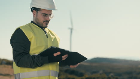 A-focused-young-engineer-in-a-white-helmet-and-reflective-vest-uses-technology-to-audit-wind-turbines-on-a-sunny-day,-emphasizing-the-importance-of-clean-energy-for-the-planet