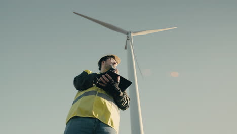 A-young-and-focused-Caucasian-male-engineer-in-a-white-helmet-uses-technology-on-a-tablet-to-audit-wind-turbines-on-a-sunny-day,-promoting-the-use-of-clean-energy-for-a-sustainable-future