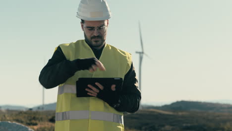 Promoting-sustainable-energy-sources,-a-professional-engineer-in-a-white-helmet-and-reflective-vest-uses-a-tablet-to-audit-wind-turbines-on-a-sunny-day-in-a-field-of-renewable-energy-generators