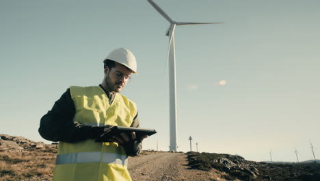 A-renewable-energy-engineer-in-a-white-helmet-uses-a-tablet-to-inspect-wind-turbines-on-a-sunny-day,-promoting-the-use-of-clean-energy-for-a-greener-future