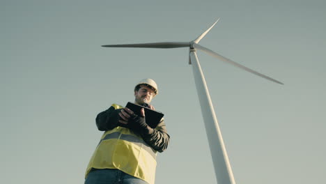 A-professional-Caucasian-engineer-in-a-reflective-vest-and-white-helmet-uses-a-tablet-to-audit-wind-turbines-in-a-field,-symbolizing-the-growth-of-renewable-energy
