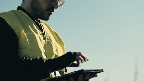 Advancing-sustainable-energy,-a-professional-engineer-in-a-white-helmet-and-reflective-vest-uses-a-tablet-to-audit-wind-turbines-on-a-sunny-day-in-a-field-of-clean-energy-generators