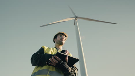 A-professional-engineer-in-a-reflective-vest-and-white-security-helmet-uses-a-tablet-to-inspect-wind-turbines,-emphasizing-the-role-of-renewable-energy-in-protecting-the-environment