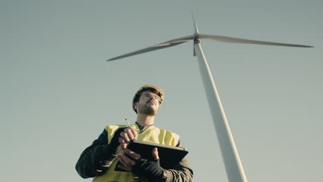 Working-to-promote-eco-friendly-energy,-an-engineer-in-a-white-helmet-and-reflective-vest-uses-technology-to-audit-wind-turbines-in-a-field-of-renewable-energy-generators-on-a-sunny-day