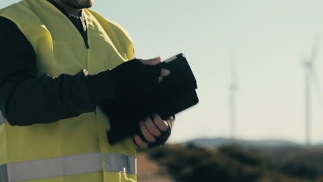 Advancing-clean-energy,-a-professional-engineer-wears-a-reflective-vest-while-using-technology-software-on-a-tablet-to-audit-wind-turbines-on-a-sunny-day-in-a-field-of-renewable-energy-generators