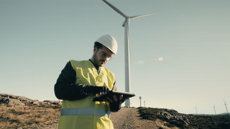 A-professional-focused-engineer-in-a-white-helmet-and-reflective-vest-uses-technology-to-audit-wind-turbines-in-a-field-of-renewable-energy-generators-on-a-sunny-day,-promoting-eco-friendly-energy