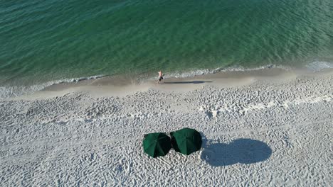 Drone-view-of-man-running-on-white-sandy-beaches-of-the-Gulf-of-Mexico-with-clear-emerald-eater-and-two-green-umbrellas-on-the-beach