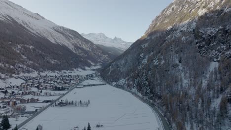 Sideways-drone-aerial-of-red-train-in-Switzerland-riding-through-stunning-snowy-winter-landscape-next-to-flowing-river-with-alpine-mountains-panorama