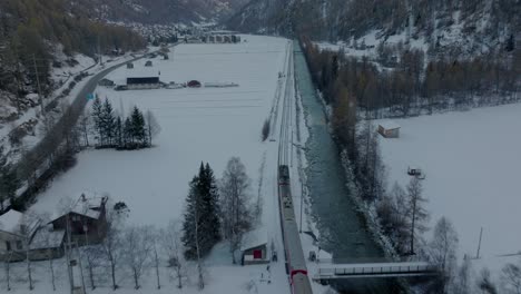 Luftdrohnenvideo-Eines-Roten-Zuges-In-Der-Schweiz,-Der-Durch-Eine-Atemberaubende-Verschneite-Winterlandschaft-Neben-Einem-Fließenden-Fluss-Mit-Alpinem-Bergpanorama-Fährt