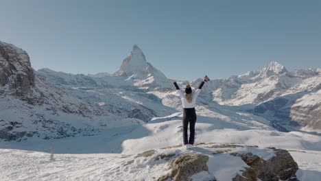 Happy-young-woman-on-snowy-rock-turning-and-raising-arms-in-breathtaking-Swiss-Alps-mountain-panorama