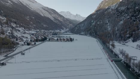 Luftdrohnenaufnahme-Eines-Roten-Zuges-In-Der-Schweiz,-Der-Durch-Eine-Atemberaubende-Verschneite-Winterlandschaft-Neben-Einem-Fließenden-Fluss-Mit-Alpenpanorama-Und-Einem-Niedlichen-Kleinen-Schweizer-Bergdorf-Fährt