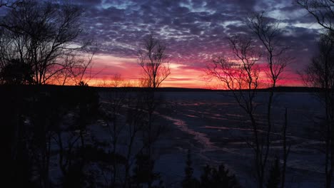 Aerial,-dramatic-red-and-orange-sunset-at-dawn-at-a-frozen-lake-during-winter