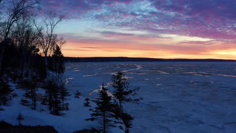 Aerial,-dramatic-orange-and-pink-clouds-at-dawn-at-a-frozen-lake-during-winter
