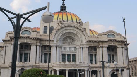 A-full-shot-with-tracking-of-the-Palacio-de-Bellas-Artes-in-Mexico-City,-with-street-lamps-in-the-foreground,-on-a-clear-day-with-blue-sky