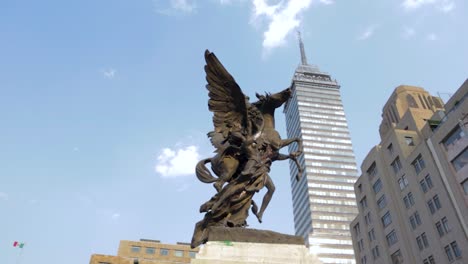 A-wide-slow-motion-shot-with-dolly-in-of-a-pegasus-and-an-angel-monument-in-the-foreground,-and-the-Latinoamericana-tower-from-Mexico-City-in-the-background-on-a-clear-blue-day