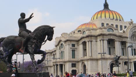 A-full-shot-with-tracking-of-a-monument-of-a-man-on-a-horse-with-the-Palacio-de-Bellas-Artes-in-Mexico-City,-with-some-people-walking-around-on-a-clear-afternoon