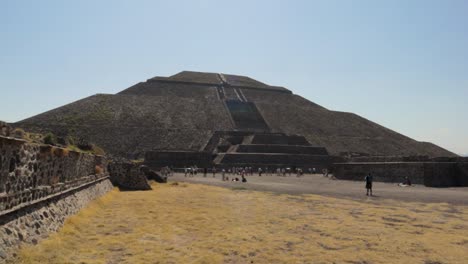 A-panning-wide-and-slow-motion-shot-of-the-Pyramid-of-the-Sun-at-the-Teotihuacan-archaeological-site-in-Mexico,-with-some-tourists-walking-around-and-the-blue-sky-on-a-clear-and-sunny-day