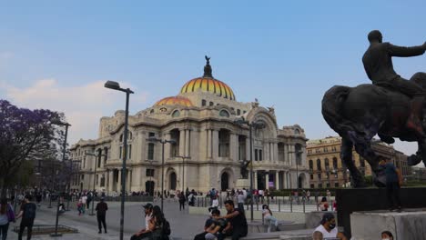 Eine-Weite-Zeitlupenaufnahme-Des-Palacio-De-Bellas-Artes-In-Mexiko-Stadt-Mit-Einigen-Herumlaufenden-Menschen-Und-Einem-Denkmal-Eines-Mannes-Auf-Einem-Pferd-An-Einem-Klaren-Nachmittag-Mit-Blauem-Himmel