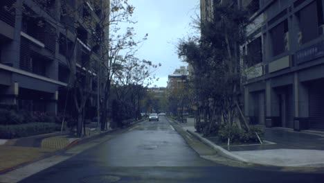 still-view-of-a-fancy-business-residential-neighborhood-area-looking-to-traffic-wet-damp-cement-road-with-a-vehicle-car-turning-into-the-underground-garage-in-gloomy-light-raining-day-with-small-trees