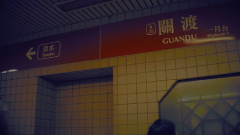New-Taipei,-Taiwan---Jan-28th-2019:-inside-look-up-view-of-Guandu-metro-station-with-poster-direction-of-Tamsui-in-Chinese-at-platform-one-underground-with-passengers-passing-by-in-Asia