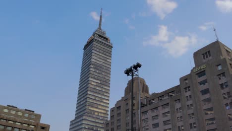 A-wide-panning-slow-motion-shot-of-the-Latinoamericana-tower-and-the-Palacio-de-Bellas-Artes-in-Mexico-City,-with-some-tourists-walking-around-on-a-clear-afternoon-with-blue-sky