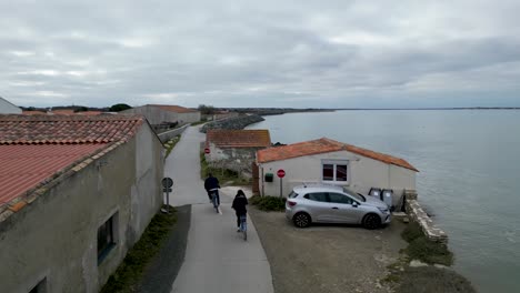 Man-overtaking-woman-in-bicycle-riding-along-the-ocean-promenade-in-Île-de-Ré-Island-Western-France-at-village-of-Loix,-Aerial-follow-behind-shot