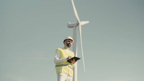 A-medium-shot-of-a-Caucasian-renewable-energy-male-engineer-using-a-tablet-to-audit-wind-turbines-in-a-field-of-clean-energy-generators