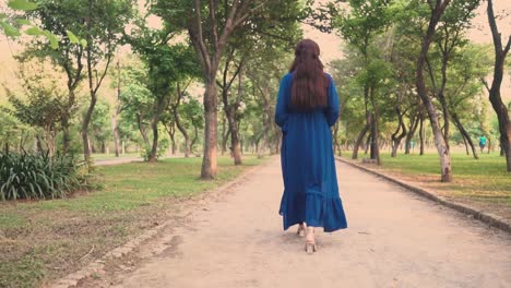 Desi-woman-is-slowly-walking-in-a-park-in-a-blue-dress-and-heels,-leisure-and-relaxing-activities-concept