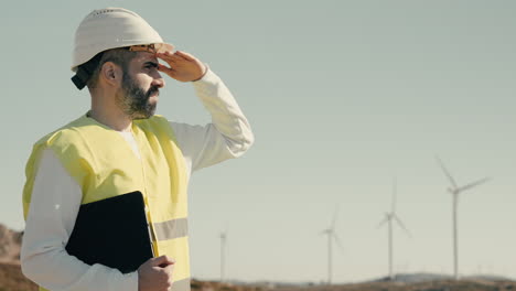 A-young-Caucasian-engineer-in-a-white-helmet-and-reflective-vest-checks-wind-turbines-on-a-sunny-day,-emphasizing-the-importance-of-renewable-energy-in-saving-our-planet