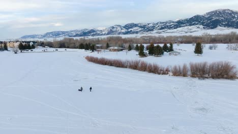 Boseman-Montana-Aerial-Winter-sunset-ice-fishermen-pulling-sled-over-snowy-suburban-park,-orbit-right-with-4k-drone-with-mountain-backdrop-at-golden-hour
