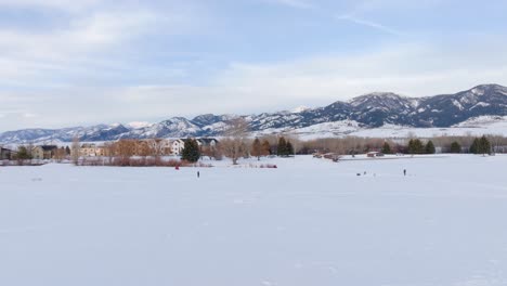 Boseman-Montana-winter-aerial-over-snowy-suburban-park-with-frozen-pond,-ice-fishermen-and-ice-skaters-with-mountain-backdrop-during-golden-hour